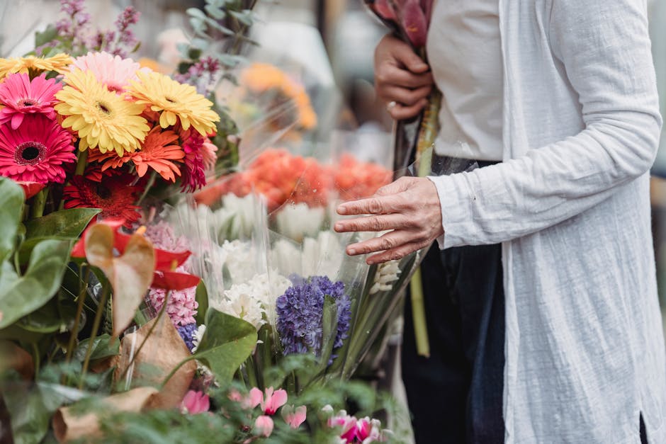 A person browsing a display of vibrant fresh flowers at a flower shop, with bouquets featuring yellow, pink, red, purple, and white blooms. The arrangement includes gerbera daisies, hyacinths, and oth
