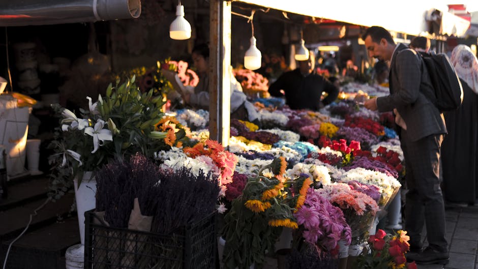 A flower stall at an outdoor market displays a vibrant variety of fresh floral arrangements, including chrysanthemums, roses, and lilies in bright pinks, whites, yellows, and purples, wrapped in clear