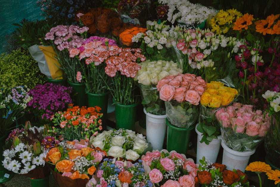 A colorful display of fresh floral bouquets and potted plants arranged on a market stall, featuring a variety of flowers such as pink and peach roses, white and yellow daisies, purple chrysanthemums,