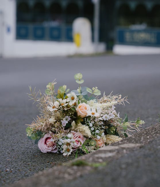 A delicate bouquet of fresh flowers rests on the pavement near the edge of a curb, arranged in a loose, natural style. The bouquet features soft pink roses, white daisies, and small white filler flowe
