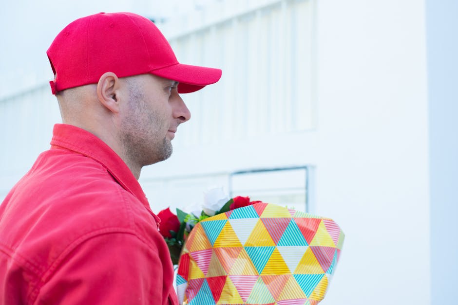 A man dressed in a red cap and jacket holds a colorful, patterned gift-wrapped bouquet of fresh flowers, featuring red roses and white blooms, with green foliage visible. The bouquet is wrapped in dec