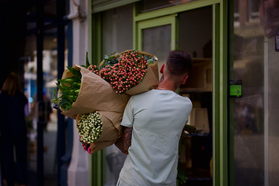 A person with short, dark hair, wearing a light-colored T-shirt, is seen from behind carrying a large bouquet of fresh flowers wrapped in brown paper. The bouquet includes clusters of pink and white b