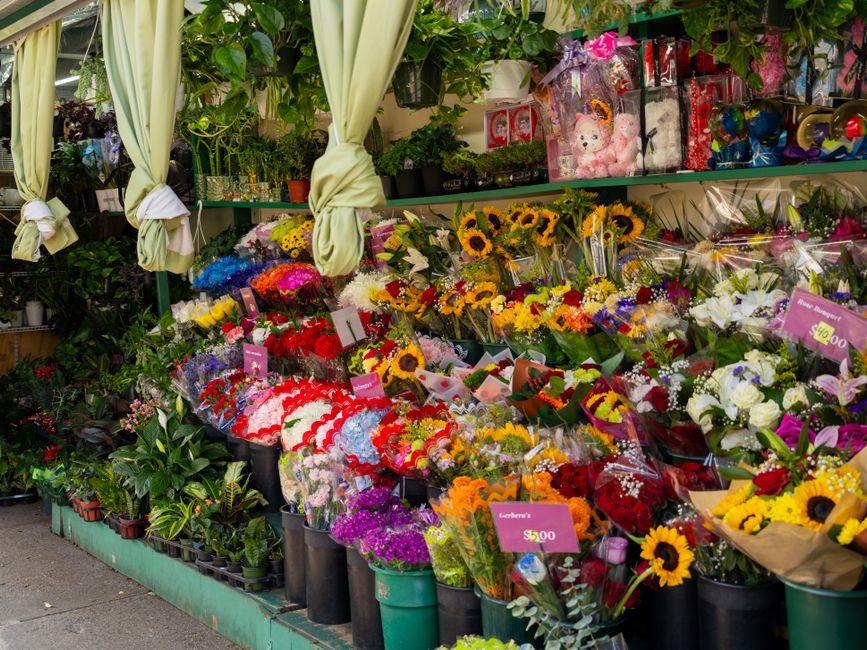 A vibrant floral display outside a flower shop featuring an assortment of fresh flower bouquets arranged in black buckets, including sunflowers, roses, lilies, and mixed seasonal blooms in bright yell