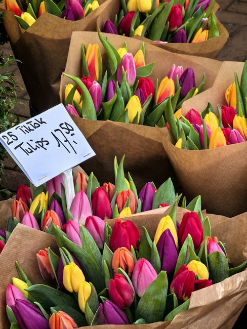 Multiple bouquets of fresh tulips wrapped in brown kraft paper, arranged outdoors on a table. The tulips feature vibrant pink, yellow, orange, purple, and red petals with green stems and leaves. Each 
