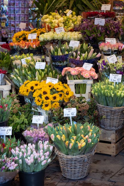 A vibrant display of fresh flowers at a floral market, featuring yellow sunflowers, pink and purple tulips, white calla lilies, and purple statice arranged in baskets and pots. The flowers are in vari