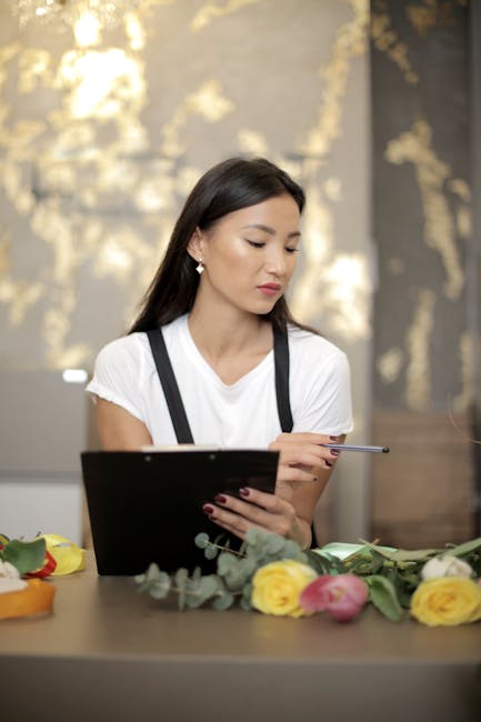 A young woman with long dark hair, wearing a white t-shirt and black suspenders, sits at a table with a tablet in her left hand and a pen in her right hand. In front of her, there is a colorful floral