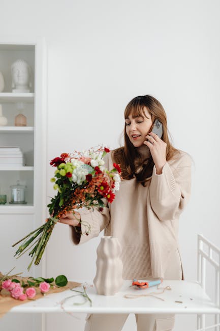 A young woman with light brown hair and bangs, wearing a cream-colored sweater, is standing in a bright, white room with a minimalist interior. She is holding a large bouquet of fresh flowers, includi