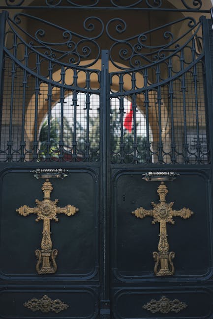 A decorative wrought iron gate painted in dark blue with intricate scrollwork at the top and golden ornate crosses affixed to the lower panels. The gate is set within a stone or brick archway, and beh