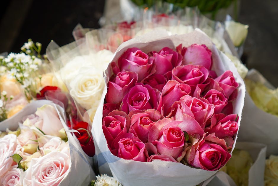 A bouquet of vibrant pink roses with tightly packed petals and soft green stems, arranged in a white wrapping paper and ribbon, placed among other floral bouquets featuring pastel pinks, whites, and h