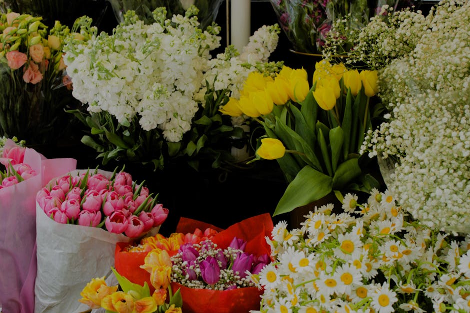 A vibrant display of fresh floral arrangements featuring bright yellow tulips in a clear glass vase, surrounded by clusters of white daisies with yellow centers, delicate white baby's breath, and soft
